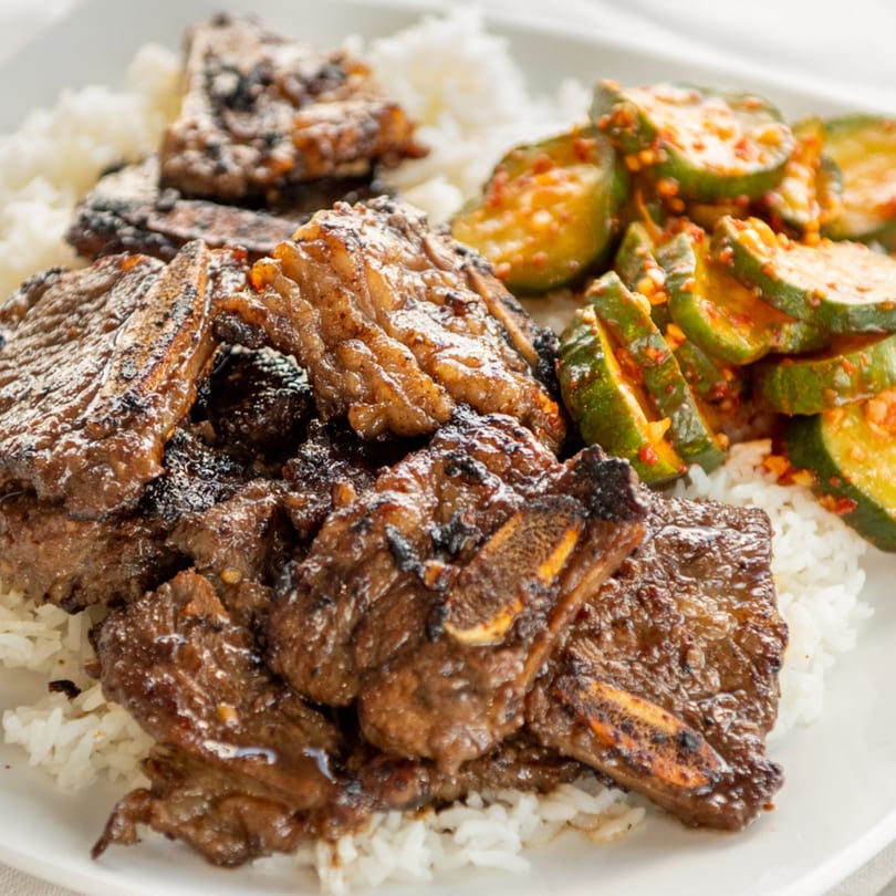 Grilled short ribs with white rice and spicy cucumber salad on a white plate.